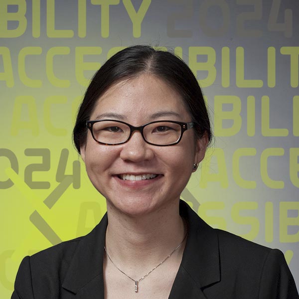 Headshot of Camille in a suit and wearing black-rimmed glasses standing against a grey background